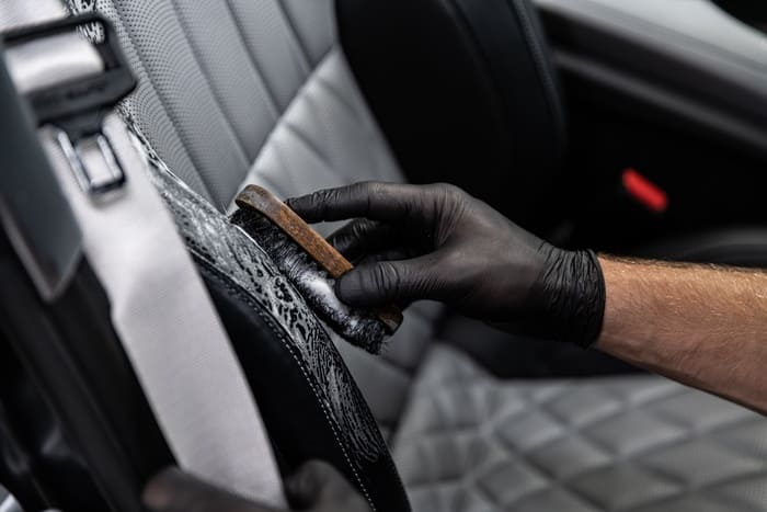 Person wearing black gloves cleaning a car's leather steering wheel with a brush and foam.