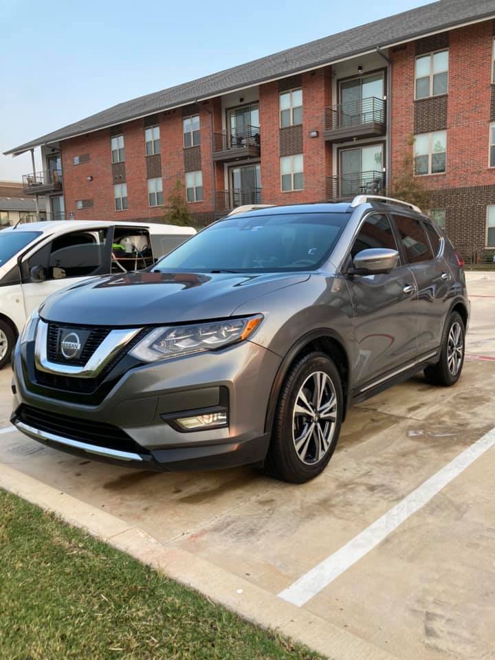 Gray Nissan SUV parked in an outdoor parking lot beside a white van, with an apartment building in the background.